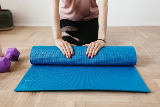 Female preparing for workout by rolling a blue yoga mat on wooden floor with dumbbells nearby.